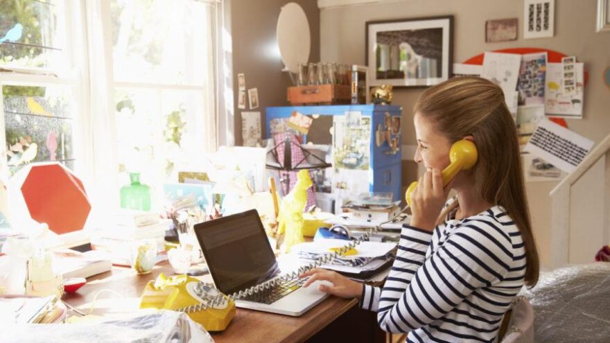 Woman on laptop running her business while working from home, with a yellow landline phone and a cluttered desk.