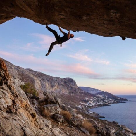 Male climber climbing along roof in cave at sunset with pink clouds against beautiful view of coast below.