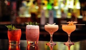 A line of cocktails, red, pink, orange and yellow, sitting on a bar with bottles of liquor blurred out behind.
