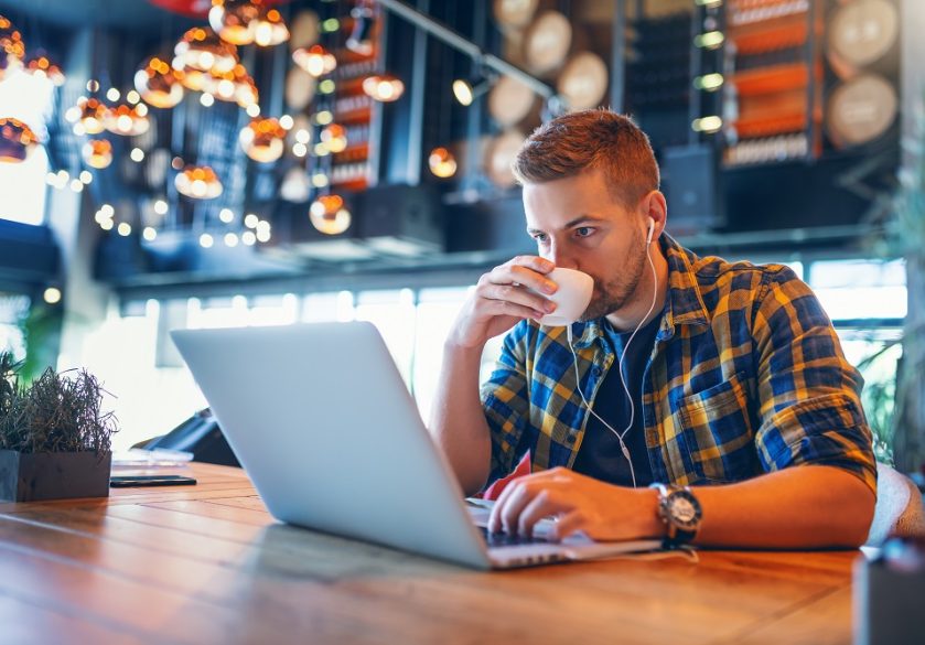 Young Caucasian blogger with earphones in ears and in plaid shirt drinking coffee and using laptop while sitting in coffee shop.