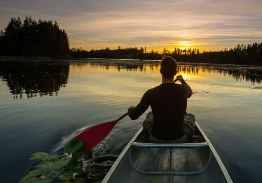 A man rowing in a canoe on a lake during the sunset, with trees in the distance.